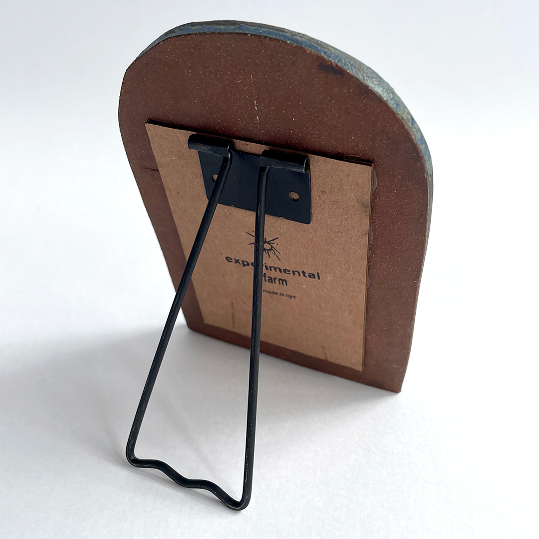 A small arched ceramic picture frame with a black metal wire support and a brown backing card printed with the Experimental Farm logo, photographed on a white background.