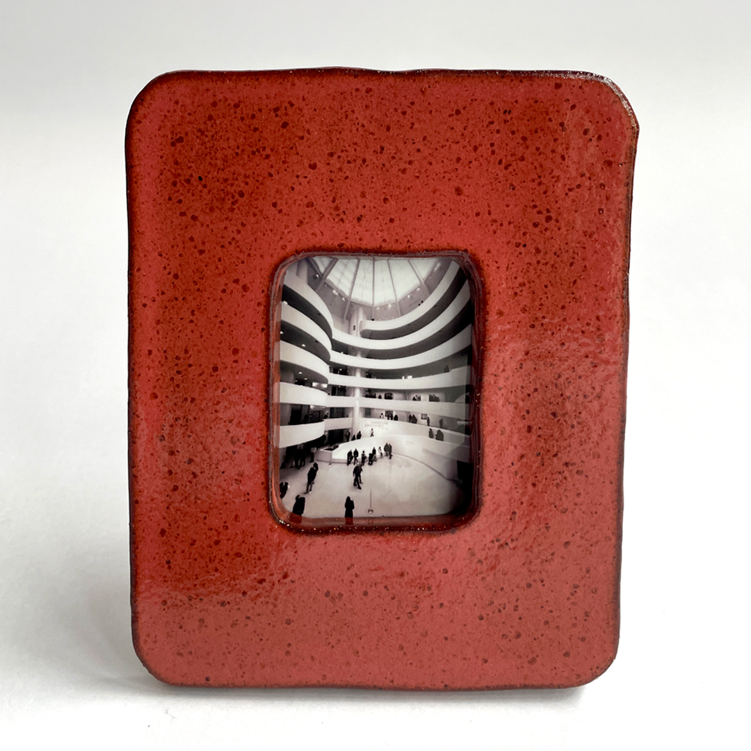 A square ceramic picture frame with a textured terracotta red glaze and a central cutout displaying a black and white photograph.