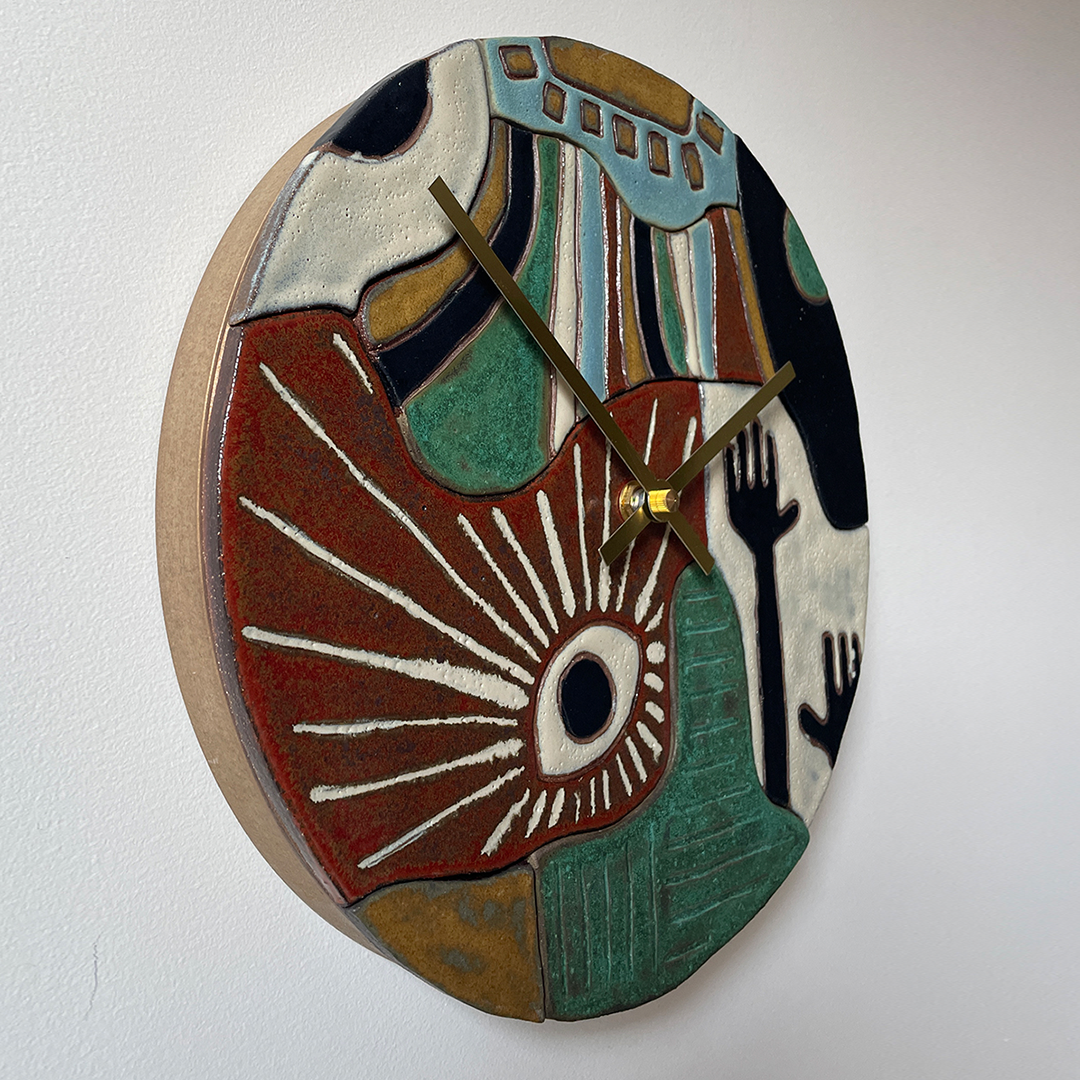 An abstract round ceramic wall clock with sculptural, raised geometric and organic shapes in terracotta red, teal green, cream, black, and tan. The clock face features a stylized eye motif and dark brown hands, mounted on a plain white wall.