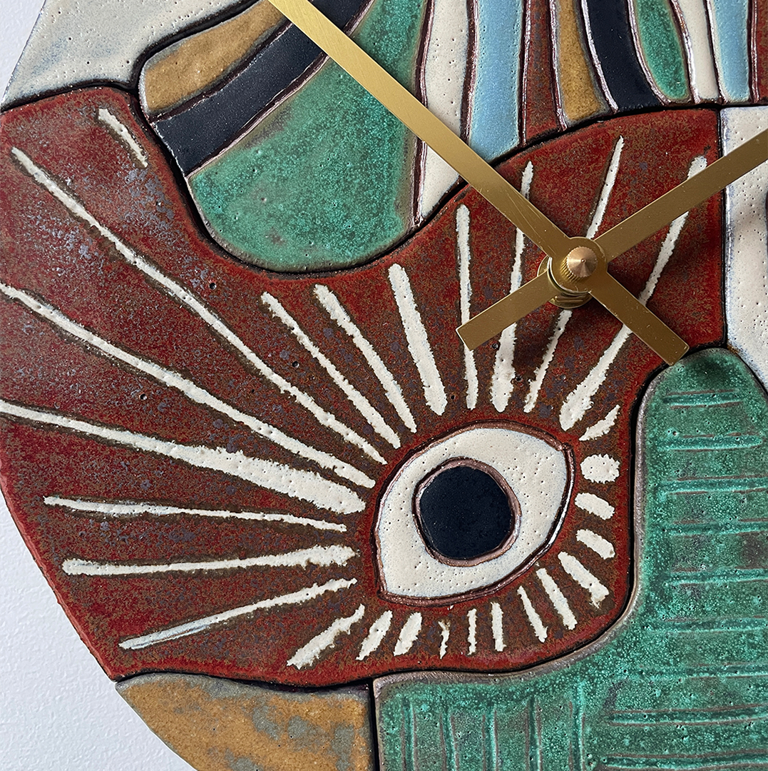 Close-up of a decorative ceramic wall clock with an abstract sculptural design. The clock face features a large stylized eye motif in red, white, black, and green, with textured ceramic-like surfaces and gold-toned clock hands.