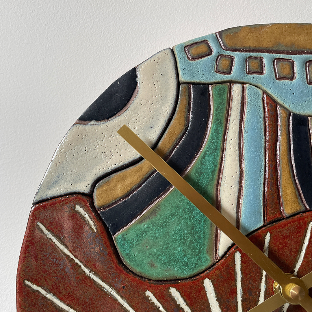 Close-up of a ceramic wall clock showing an abstract geometric design in terracotta red, mint green, light blue, cream, and dark navy, with a gold-toned hand pointing to the minute.