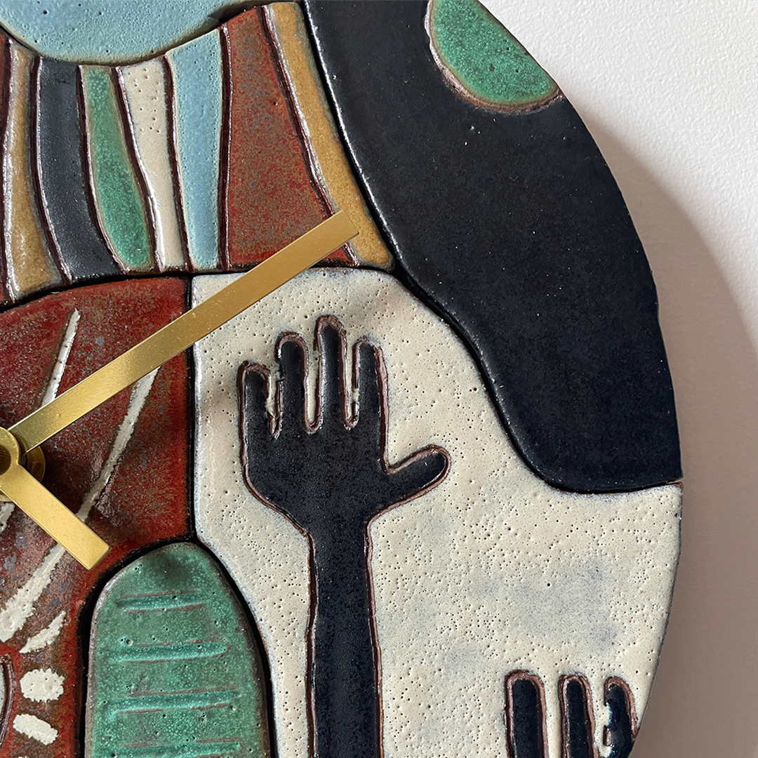 Close-up of a ceramic wall clock with an abstract, cubist-style design featuring a raised hand motif, painted in black, terracotta red, teal green, cream, and light blue on a white wall.
