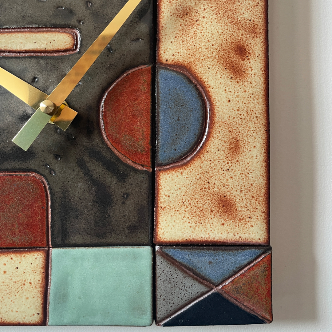 Close-up of a geometric wall clock with abstract ceramic tile faces in beige, brown, blue, green, and black, featuring gold-tone hands.