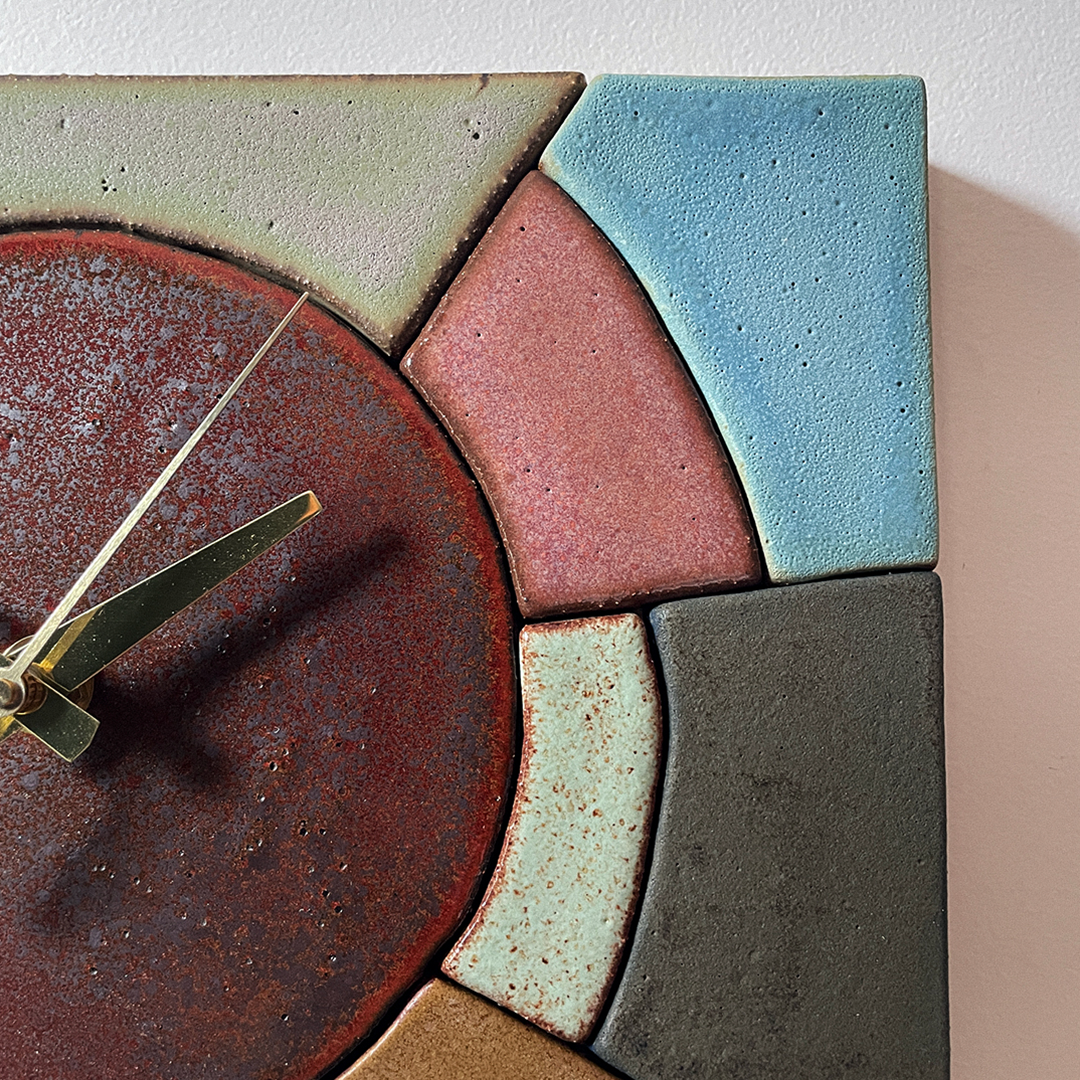 Close-up of a textured ceramic wall clock with curved and angular segments in rust red, light blue, dusty pink, charcoal gray, pale mint, and tan, with gold-tone hands against a light wall.