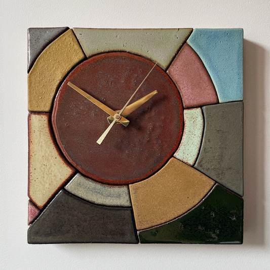 A square ceramic wall clock with a geometric mosaic design in earthy tones. The center features a brown dial with gold-tone hands, surrounded by sections in beige, gray, blue, pink, and green.
