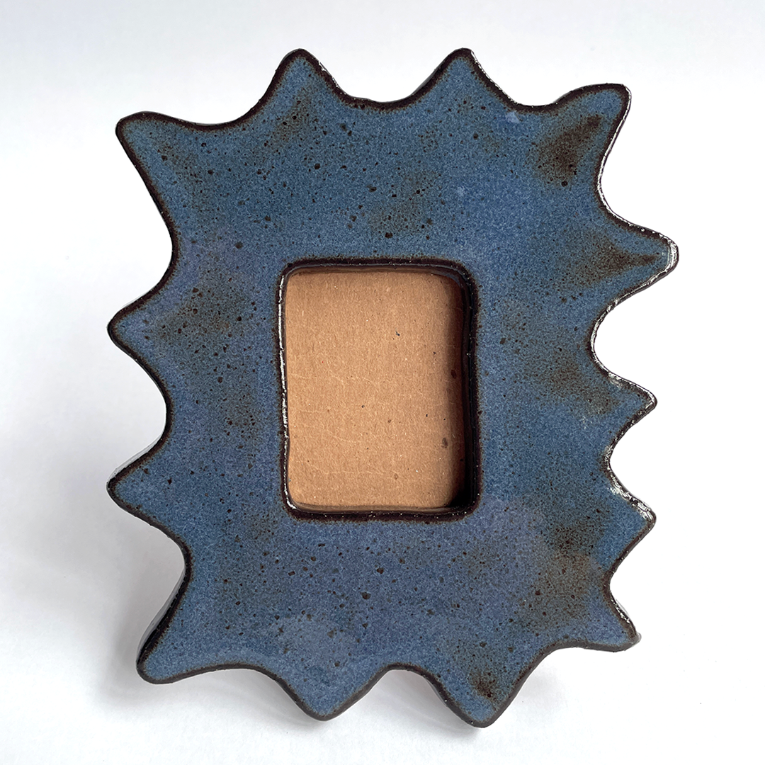 A starburst-shaped ceramic picture frame with a textured dark blue glaze and a rectangular tan center, photographed on a plain white background.