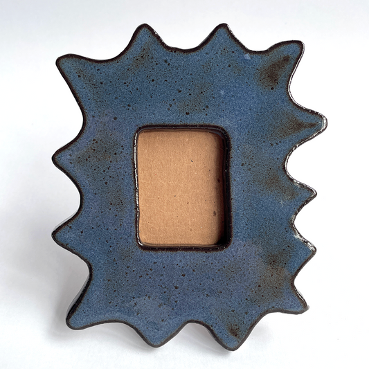 A starburst-shaped ceramic picture frame with a textured dark blue glaze and a rectangular tan center, photographed on a plain white background.
