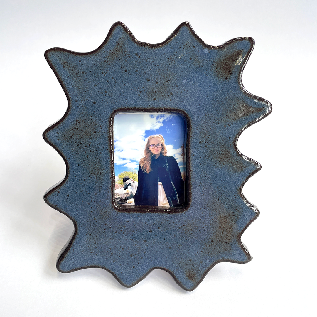 A handmade ceramic picture frame with a rustic, scalloped starburst shape in a speckled slate blue glaze. The center is open to display a custom photo of a woman, set against a plain white background.