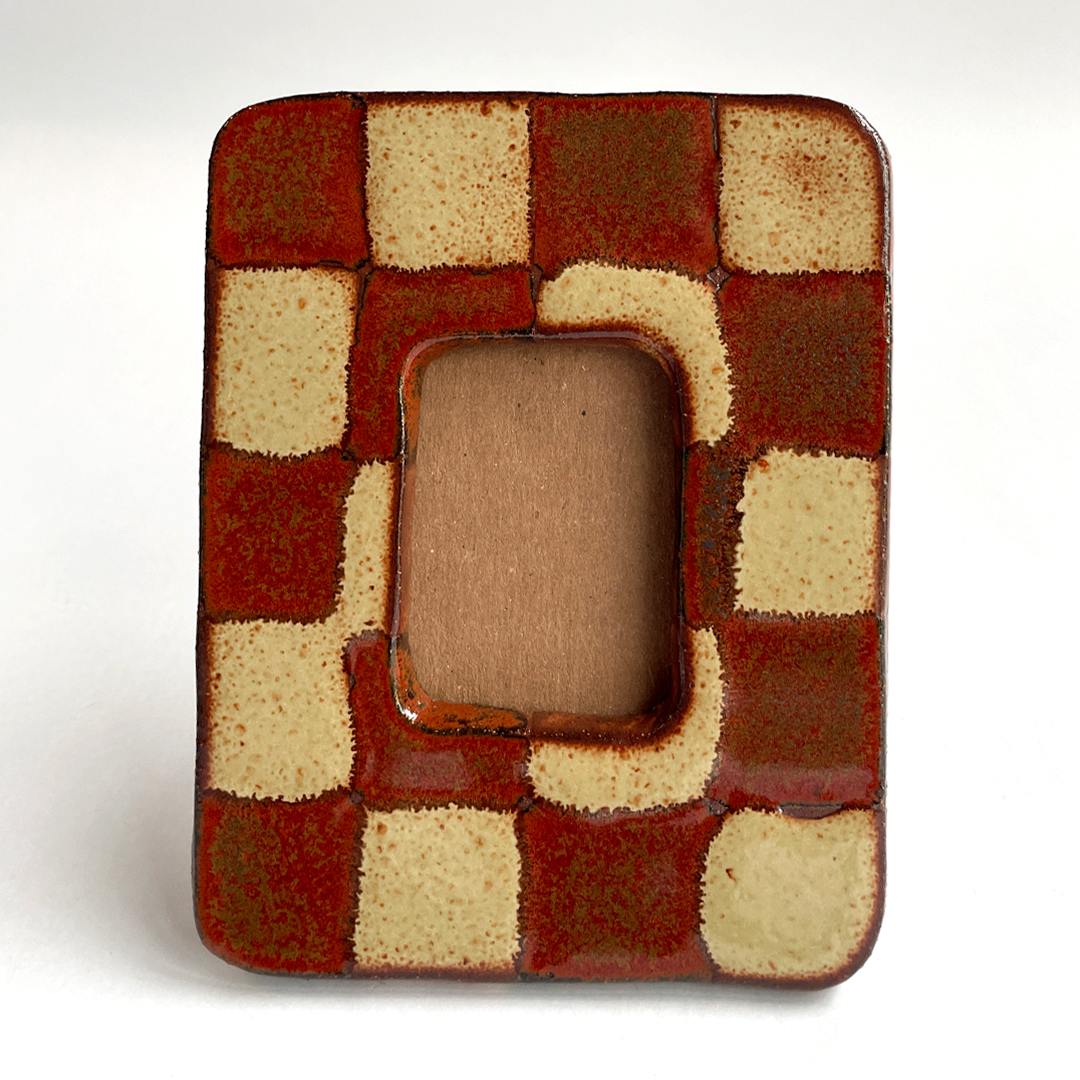 A square ceramic picture frame with a glossy checkered pattern in terracotta red and cream, featuring a central rounded rectangular cutout.