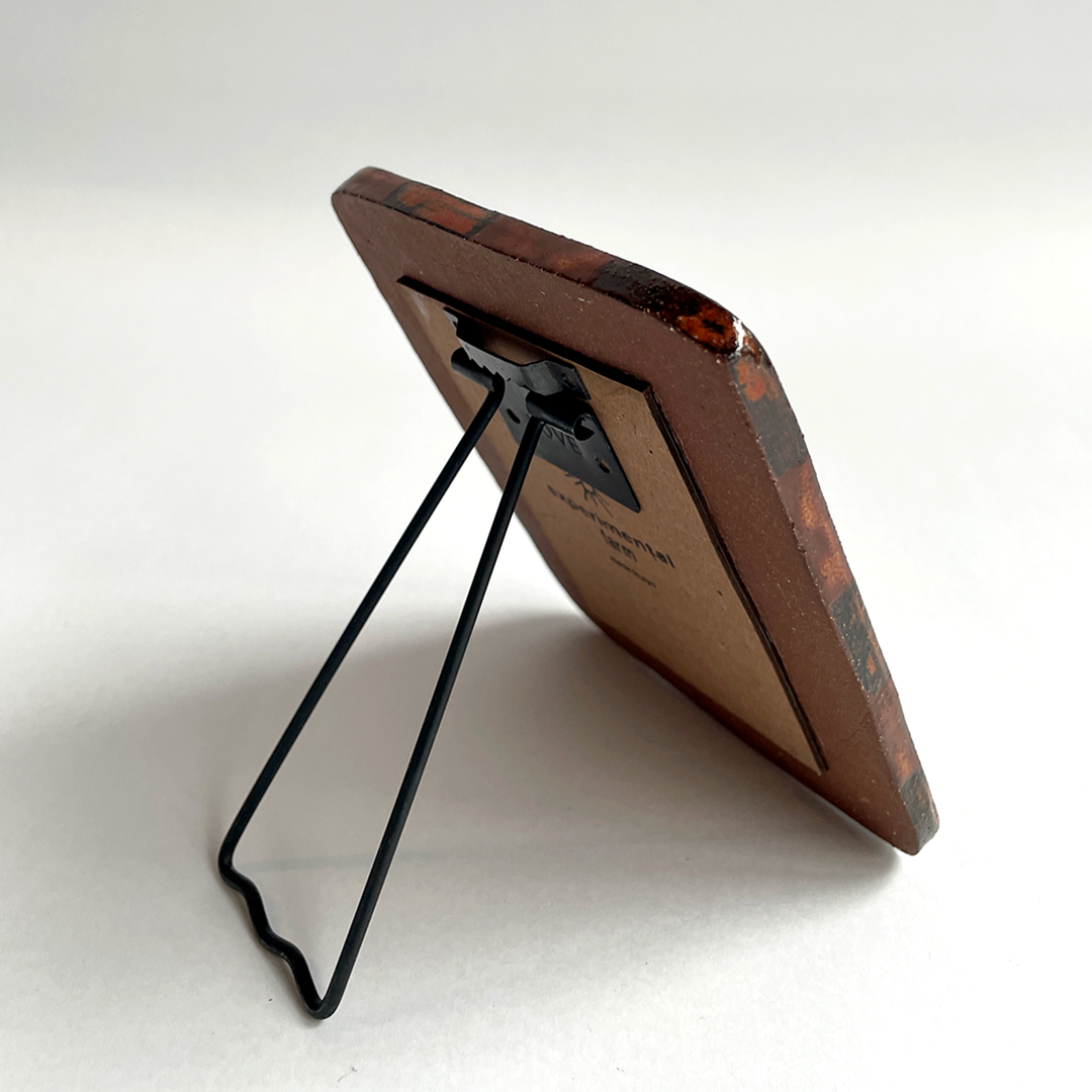 A square picture frame with a dark brown marbled patterned border and a black metal wire stand, shown from an angle on a white background.