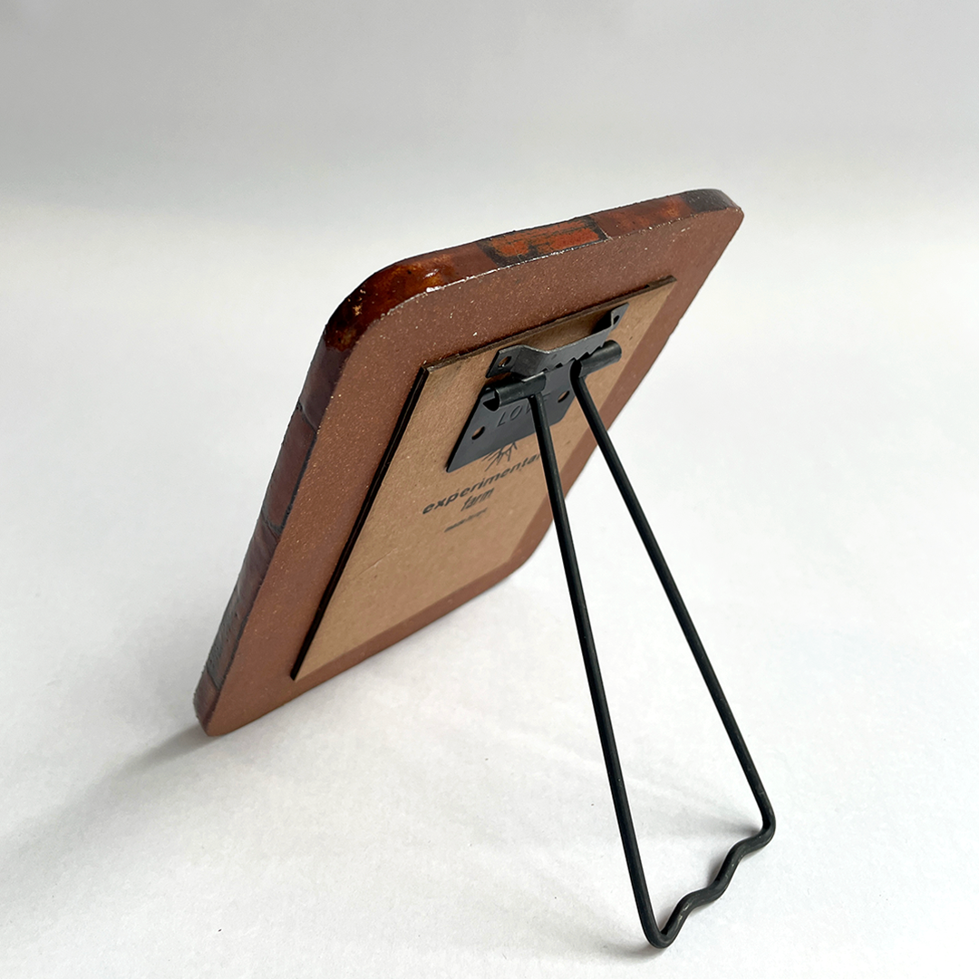 A vintage-style picture frame with a reddish-brown ceramic, a tan backing board, and a black metal wire stand, photographed on a light gray background.