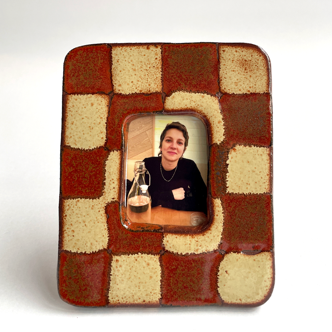 A square ceramic picture frame with a rustic checkered pattern in terracotta red and cream, featuring a central cutout holding a small portrait photo.