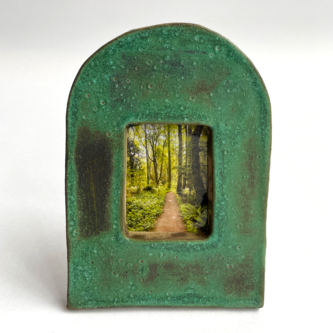 A green textured ceramic picture frame with a rounded top and a square center cutout showing a miniature forest path scene, set against a plain white background.
