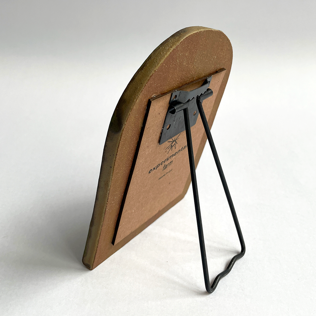 A small freestanding ceramic picture frame shown from the back on a light gray studio background. The frame has a warm brown wood or cardboard backplate with a black metal support stand and a brass-toned edge.
