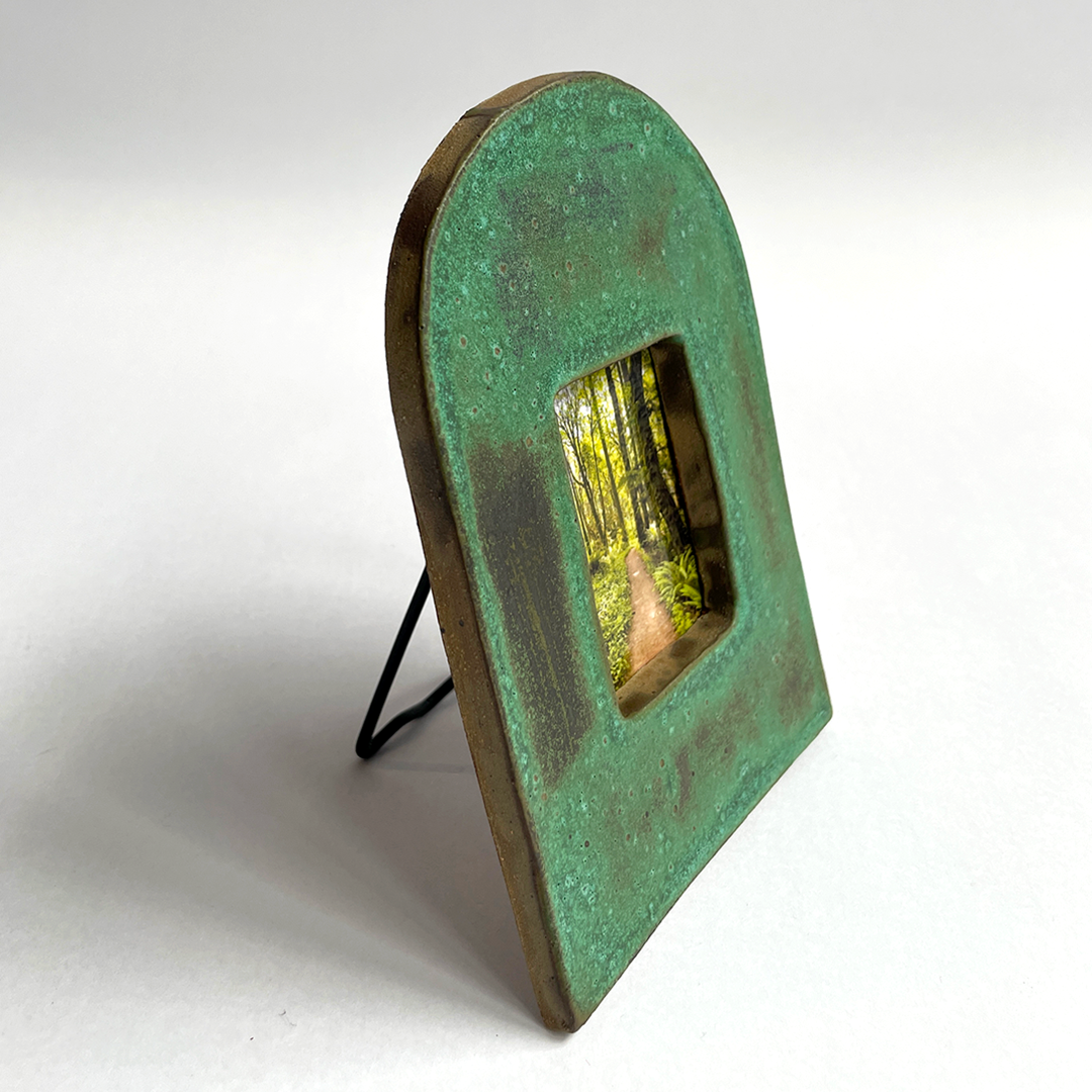 A rustic, arched ceramic picture frame with a distressed green and brown patina finish, supported by a small black wire easel stand. The center cutout displays a miniature forest scene insert.