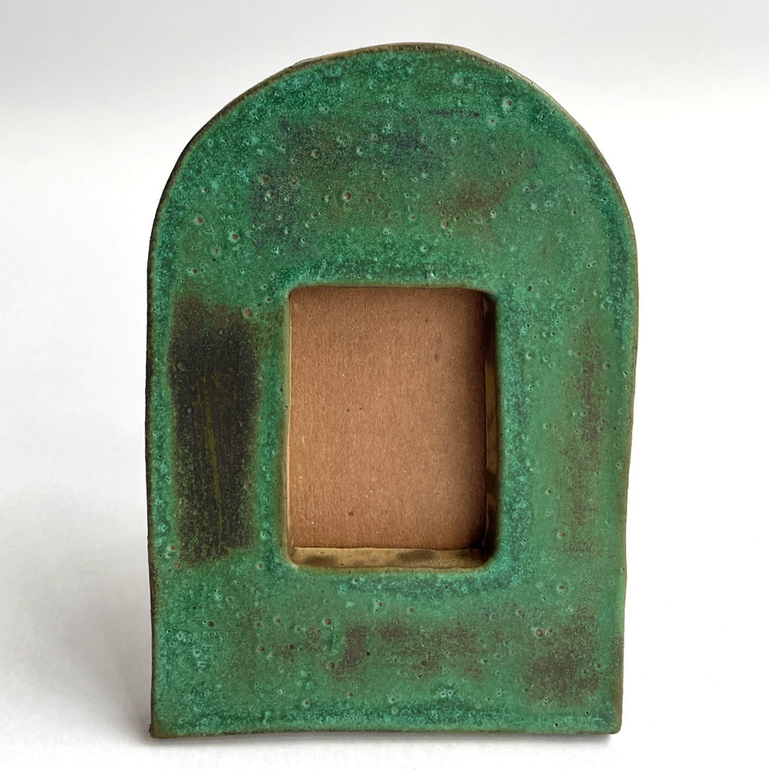A rustic green ceramic arch-shaped frame with a textured, weathered finish and a square cutout in the center showing a brown backing, set against a plain white background.