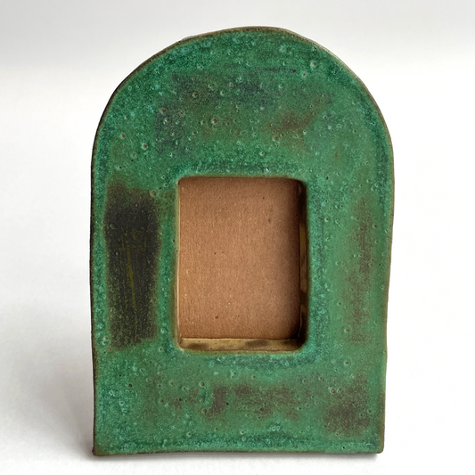 A rustic green ceramic arch-shaped frame with a textured, weathered finish and a square cutout in the center showing a brown backing, set against a plain white background.