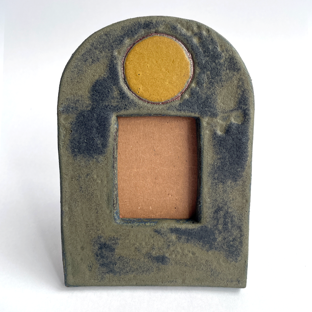 A small decorative ceramic picture frame with an arched top, a round gold-toned moon at the top, and a rectangular opening below showing a warm tan interior, photographed on a plain white background.