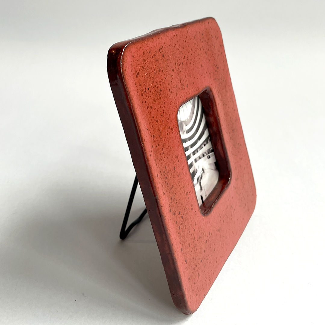 A square terracotta red picture frame with a textured glaze and a small black wire stand, shown on a white background.