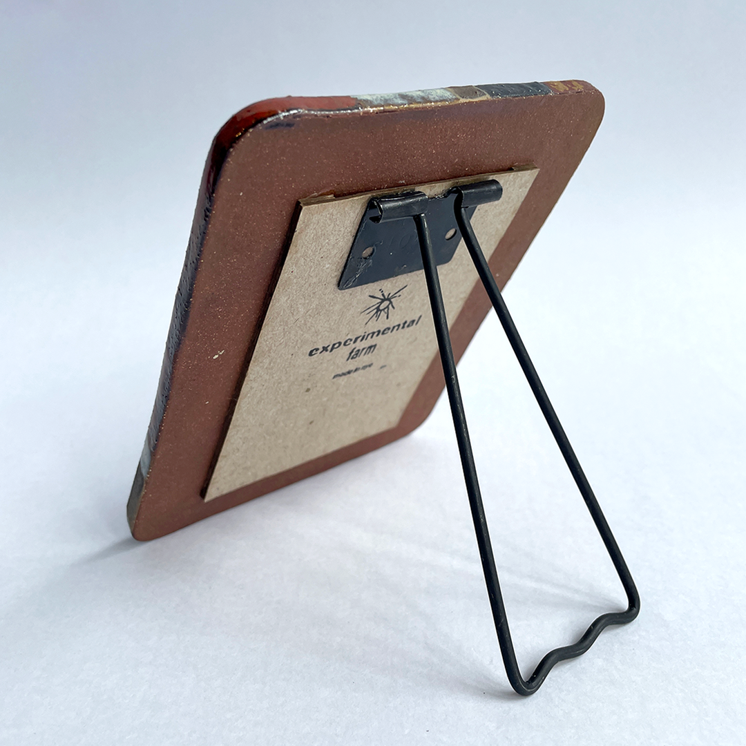 A vintage rectangular picture frame with a brown ceramic and a black metal wire stand, shown from the back on a light gray background. The backing board has 'experimental farm' printed on it.