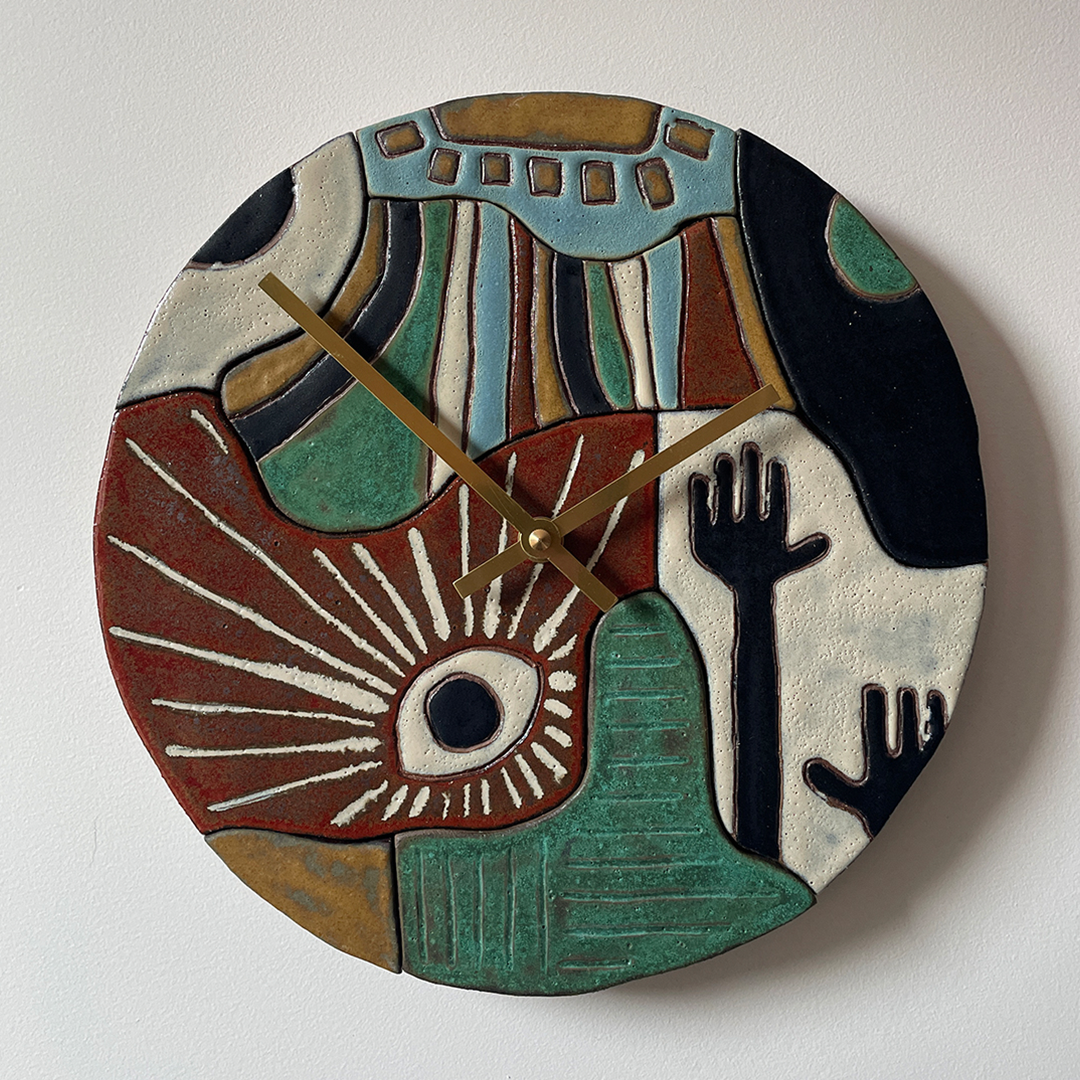 A round, hand-painted and hand-carved abstract ceramic wall clock with sculptural relief details and gold-tone hands, mounted on a plain white wall.